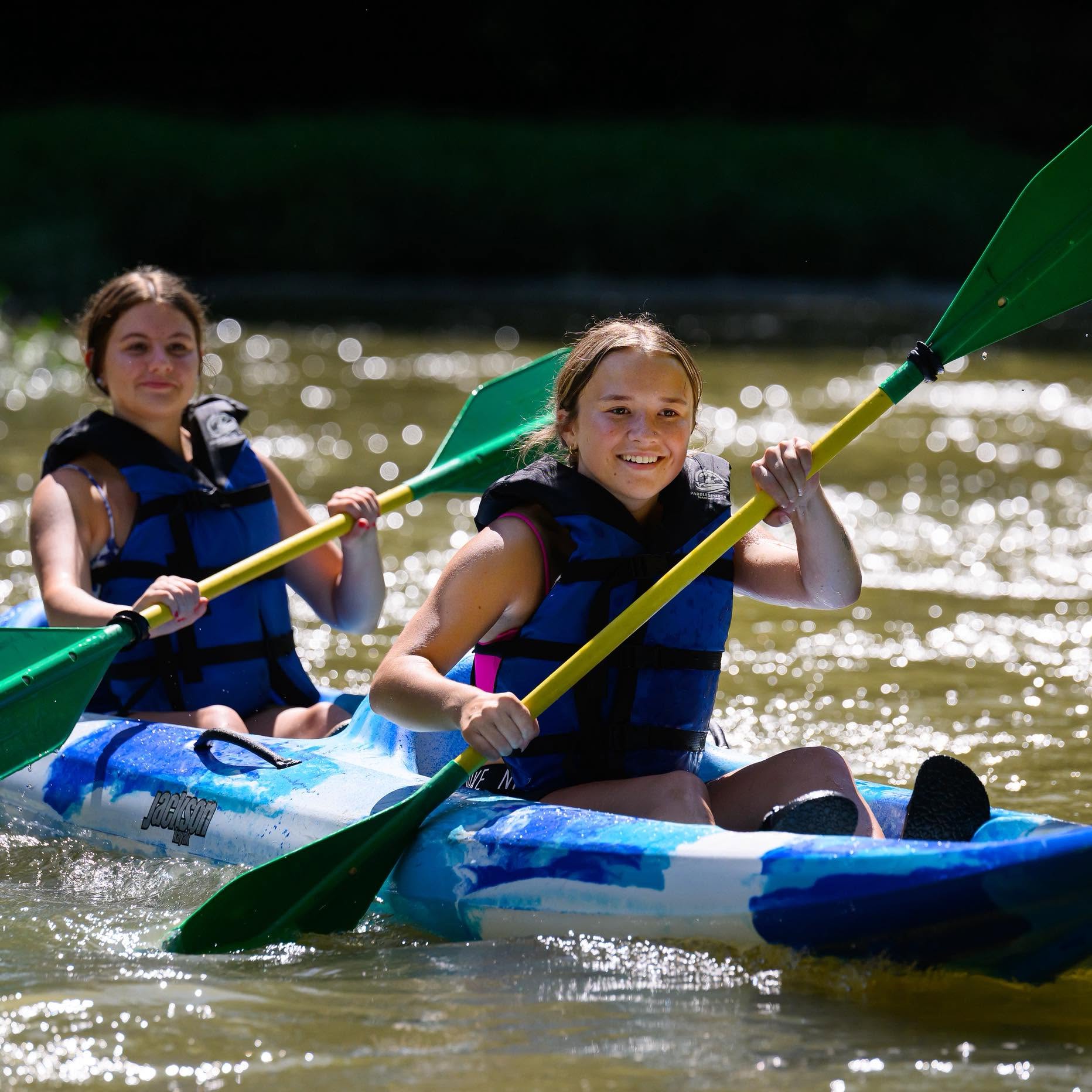 Two people wearing life jackets paddle a blue kayak on a sparkling river, holding yellow paddles under bright sunlight on a sunny day.