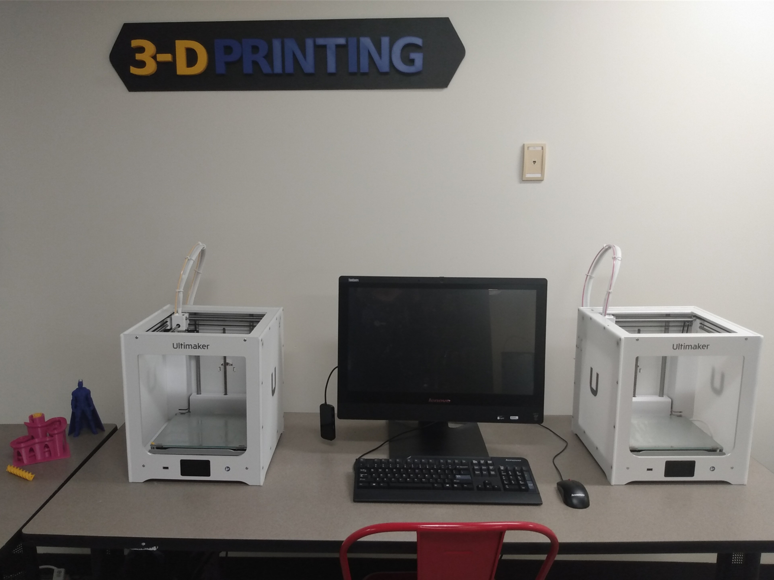 Two white Ultimaker 3D printers sit on a desk beside a central Lenovo monitor, with a 3-D PRINTING sign on the wall above.