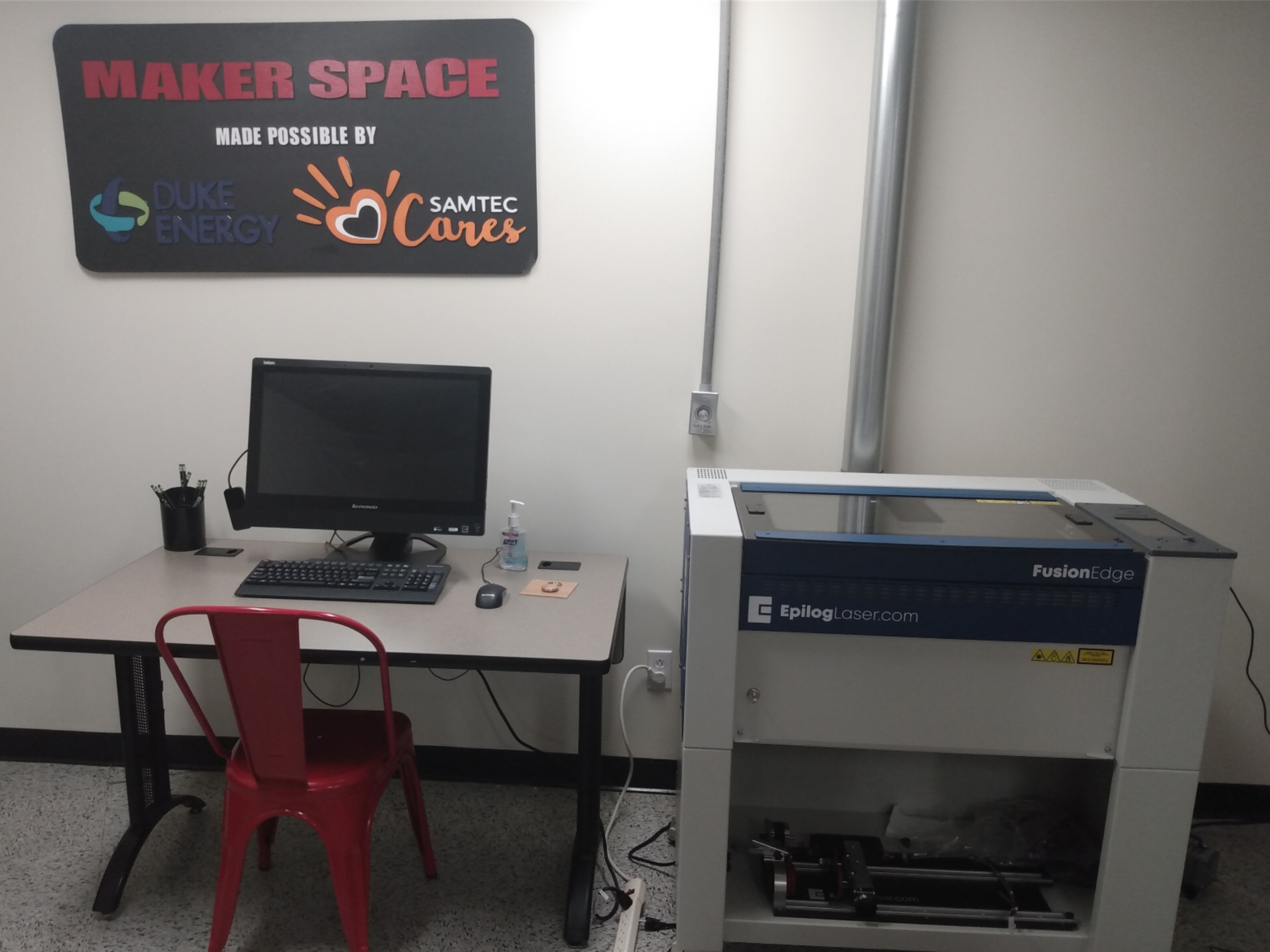 Maker space sign on the wall above a computer workstation in a workshop room with a red chair nearby.