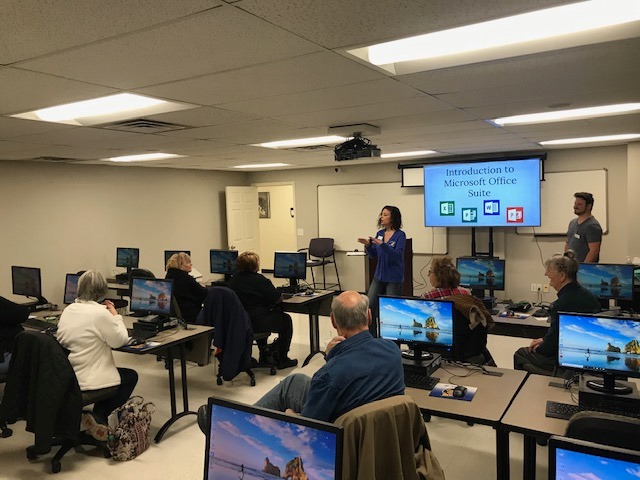 Instructor presents Microsoft Office Suite to adults in a computer-lab classroom with desktops.