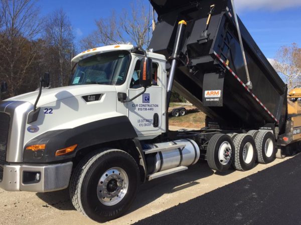 White dump truck with its black dumping bed raised, parked on a dirt road at a construction site, logos visible on the cab door.