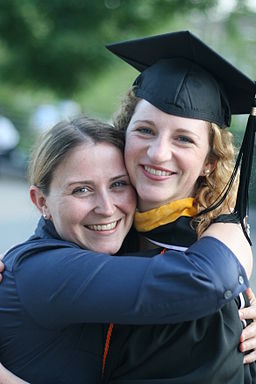 Two graduates share a hug, one in a cap and gown, smiling at a ceremony.