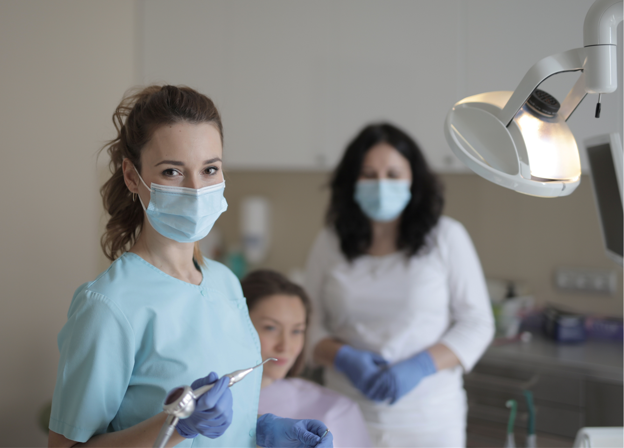 Dentist in blue scrubs and mask holding dental instruments in a clinic with an overhead light on the right side of the image.