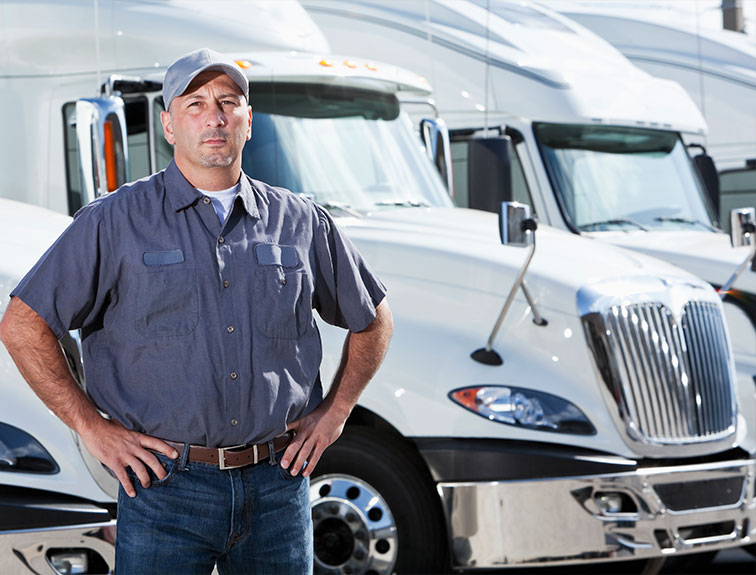 Truck driver or fleet worker stands with hands on hips in front of white semi-trucks in a yard.