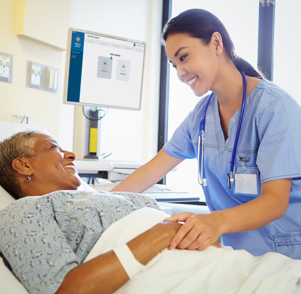 Nurse in blue scrubs leans over smiling elderly patient in bed, holding hands in a caring, bedside checkup.
